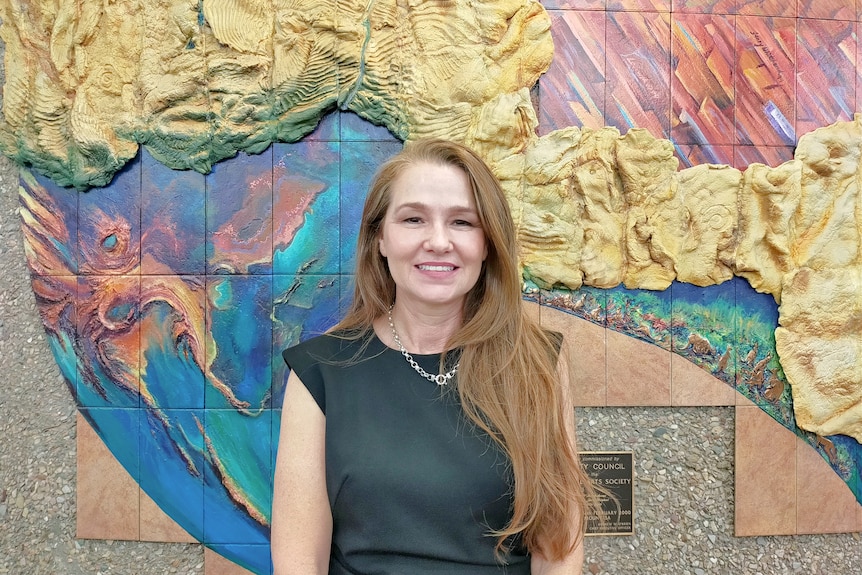 A woman with long brown hair wears a black business dress stands infront of a colourful wall