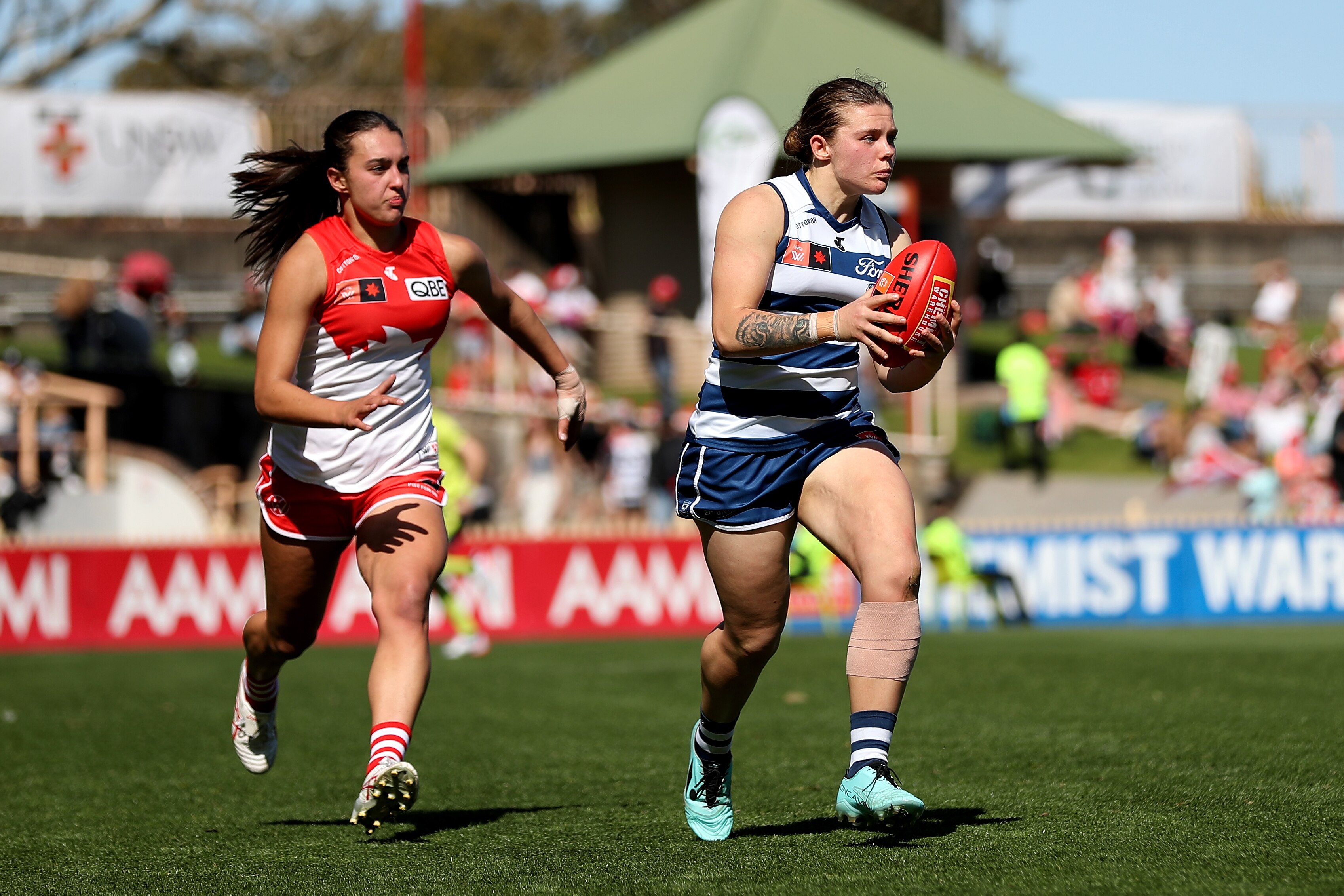 A Geelong AFLW player looks downfield as she runs with the ball as a Sydney defender trails her.