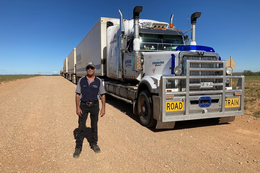 Man stands in front of large road truck on a red dirt track road. 