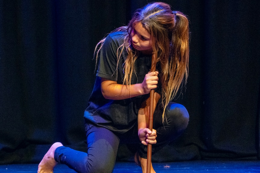 Young girl, long brown hair half tied, black tee, leggings, one knee on stage, blue lighting, holds stick and looks at someone.