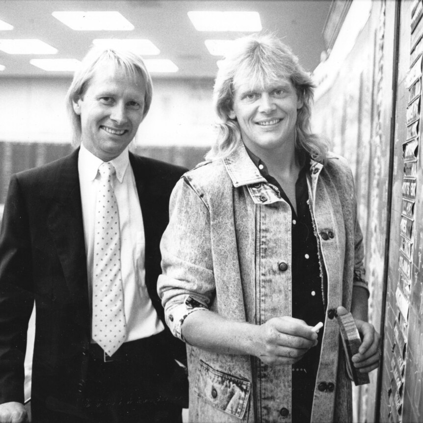A black and white photo of a young John Farnham and Glenn Wheatley smiling, Glenn in a suit, John in a denim jacket