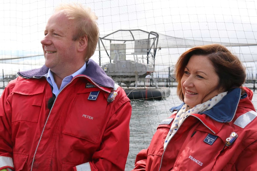Balding man and brown haired woman, both wearing red jackets and standing in front of a salmon cage.