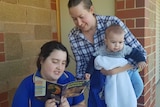 Child in blue shirt reading a book with a woman leaning over her shoulder, the woman is holding a baby