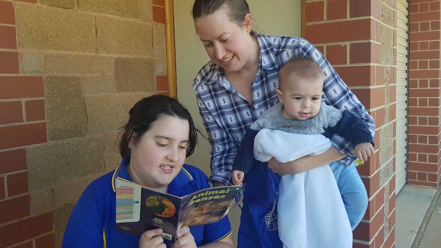 Child in blue shirt reading a book with a woman leaning over her shoulder, the woman is holding a baby