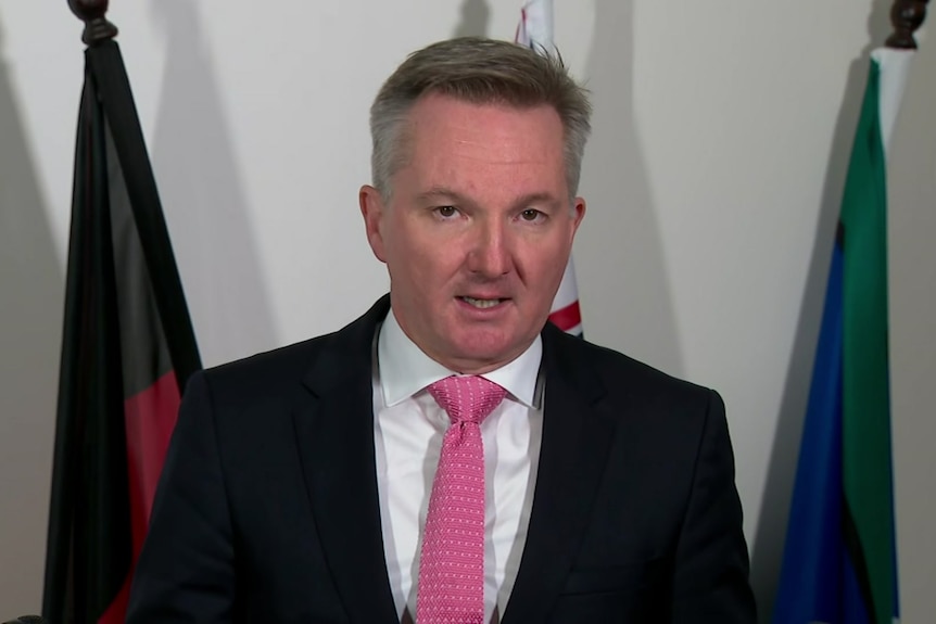 Bowen, wearing a red tie, speaks to the camera.  Behind him are three flags