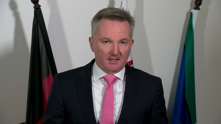 Bowen, wearing a red tie, speaks at the camera. Behind him are three flags