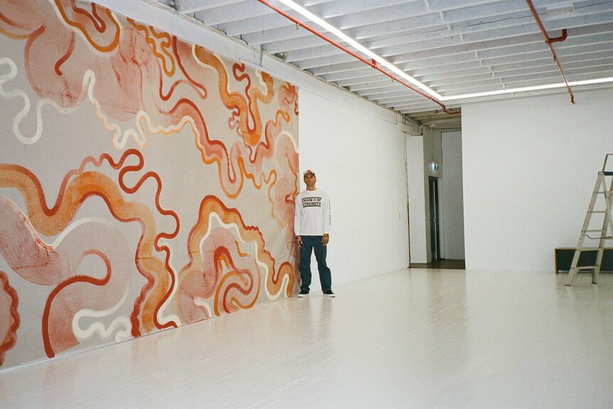 A wide shot of man leaning against a large scale orange, red and cream canvas against a white wall. 