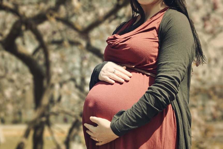 Pregnant woman stands in park and has hand on stomach.