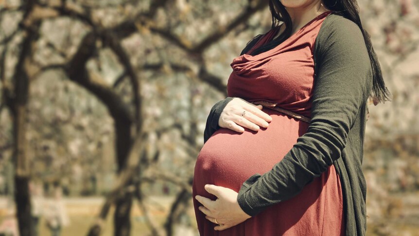 Pregnant woman stands in park and has hand on stomach.