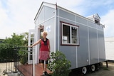 A woman stands on the front landing of a tiny home, painted grey