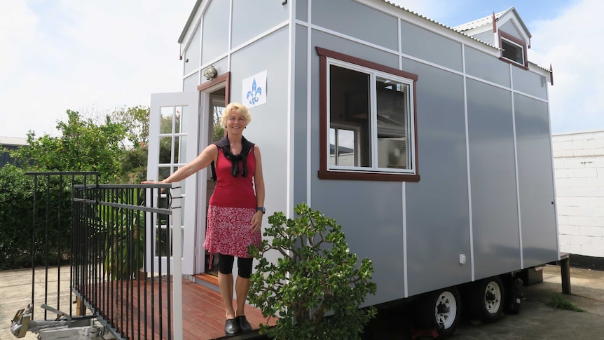 A woman, who is smiling, stands on the front wooden porch of a small demountable-like home on wheels.