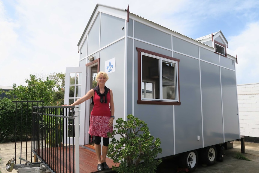 A woman, who is smiling, stands on the front wooden porch of a small demountable-like home on wheels.