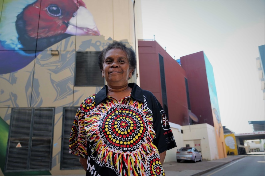 woman wearing an aboriginal print shirt standing in a shaded city lane with a finch painted on an adjacent building