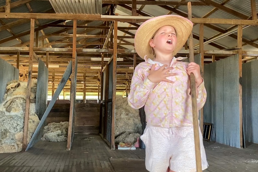 A young girl with a hat holding a wool sweeper singing in a shearing shed near Longreach, November 2022.