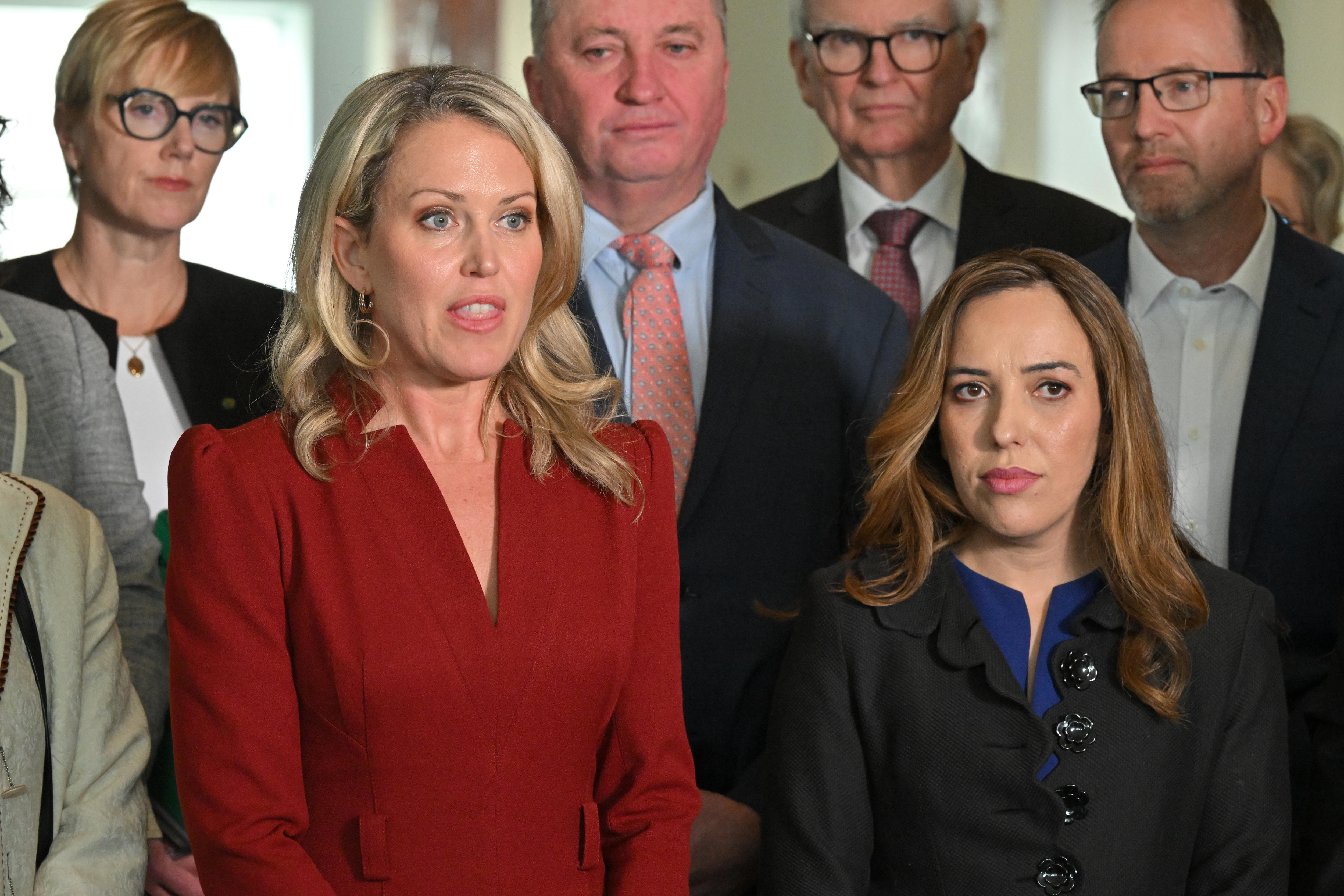 Jennifer, dressed in a red long-sleeved dress, speaks to the media as Stella stands next to her and MPs behind her.