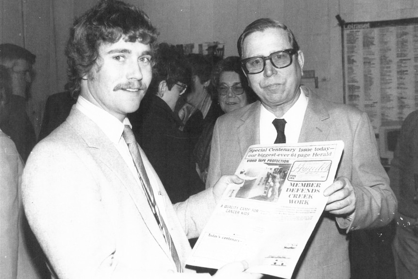 black and white photo of a ban with curly hair and moustache and his father with glasses and short hair holding a newspaper