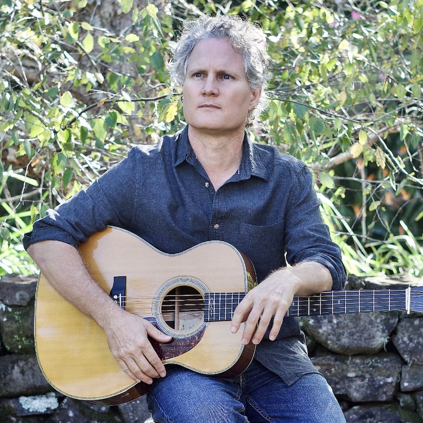 A man with curly grey hair sitting outside at a tree with guitar on his lap