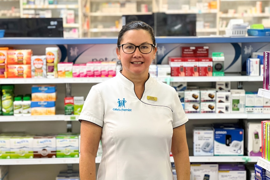 Cate Whalan wearing a white uniform with a "Cate's Pharmacy" logo, standing in front of rows of medicines on the shelves.