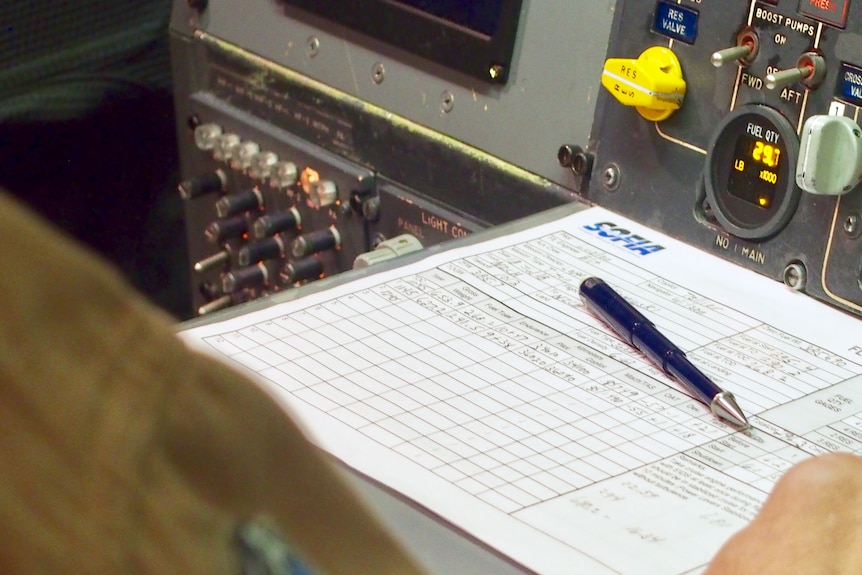 A paper chart titled "Flight engineer's fuel log" sits on a shelf next to a panel covered in switches and dials.