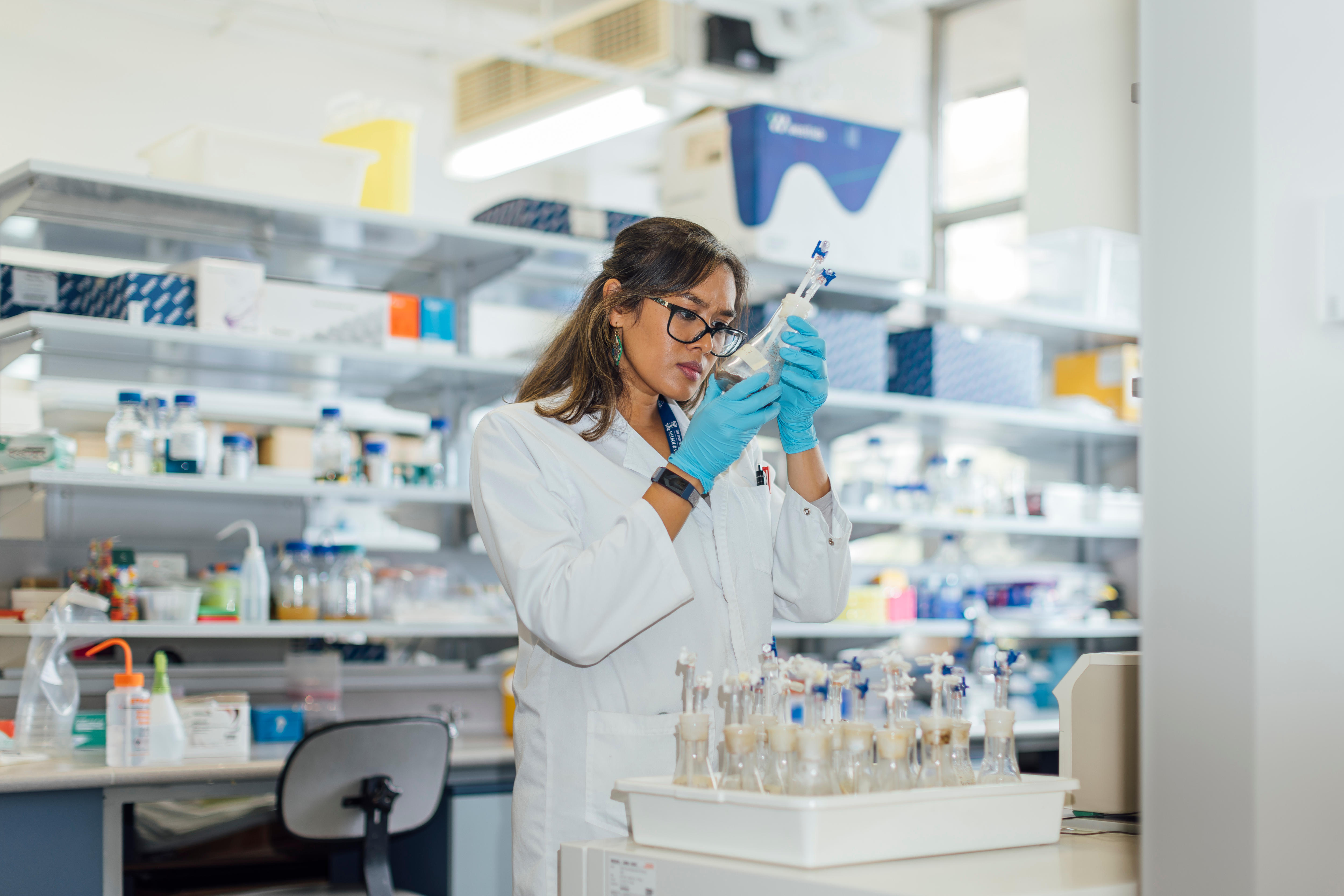 Uma mulher em um laboratório examinando amostras em um tubo de ensaio, com uma bandeja de amostras à sua frente.