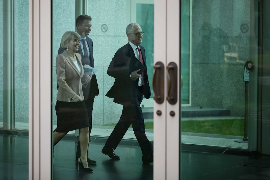 Malcolm Turnbull and Julie Bishop walk side by side, Ms Bishop smiling, down a corridor with another man