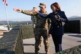 Kamala Harris stands at a barricade with an American military officer. She holds binoculars, both are pointing off to the right
