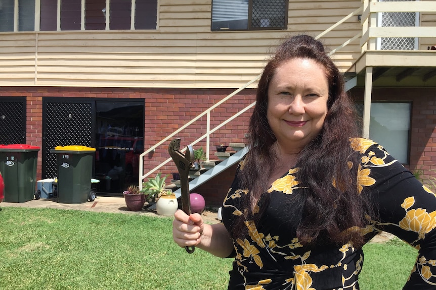 A woman holds a large spanner in front of her house.