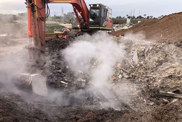 a pile of tip material with smoke coming from it,  there is heavy machinery behind it.