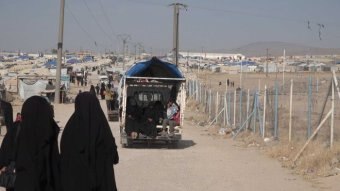 Women in black burqas walk away from the camera in front of a ute and a refugee camp.