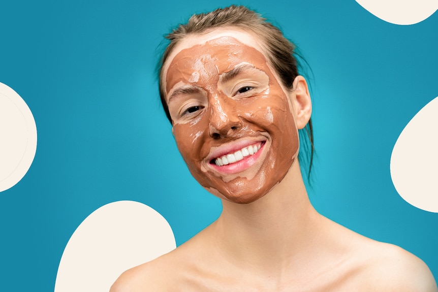 Smiling woman with a clay mask on her face, against a blue and white background.