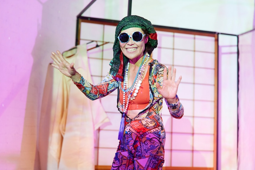 A Japanese Australian woman dressed in eclectic clothing and large sunglasses performs onstage.