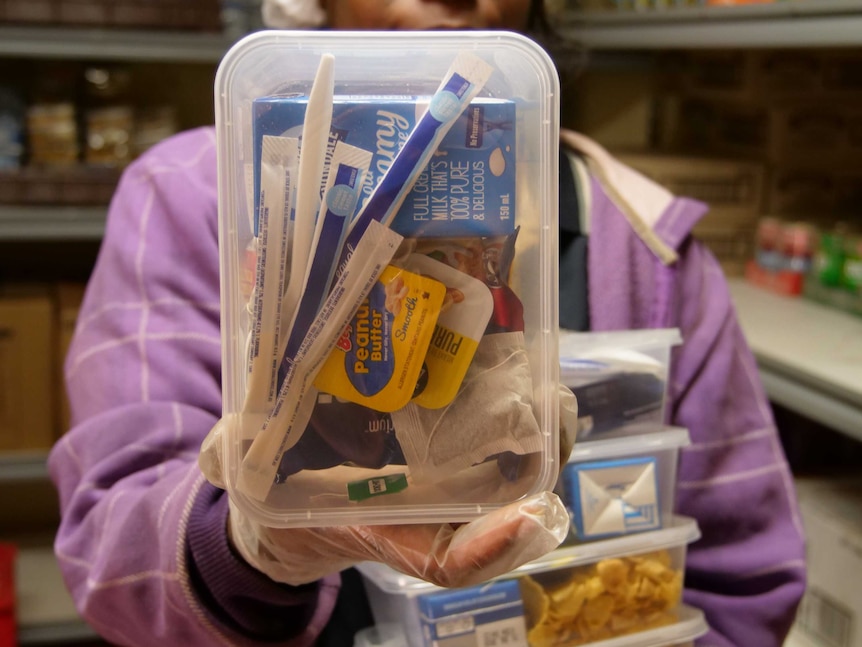 A worker holds up a transparent plastic lunch box containing sachets of sugar and spreads, teabags and long-life milk.