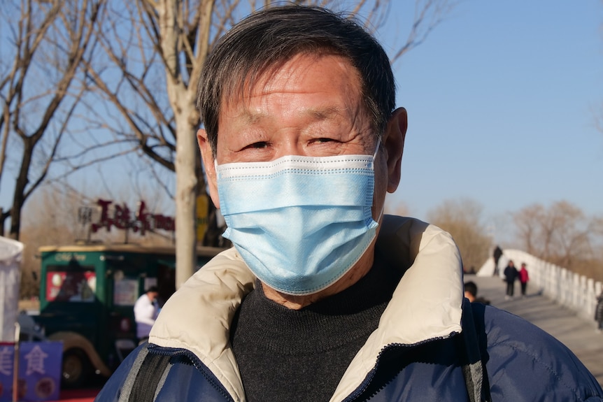 A man with black hair and wearing a face mask smiles in a park.
