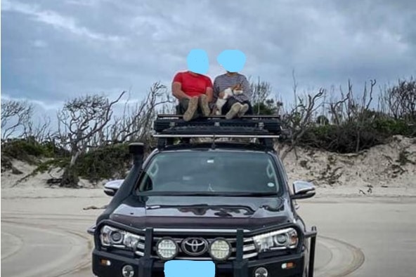 Two people with blurred faces sit on top of a FWD car on the beach, they have a ginger cat in their lap