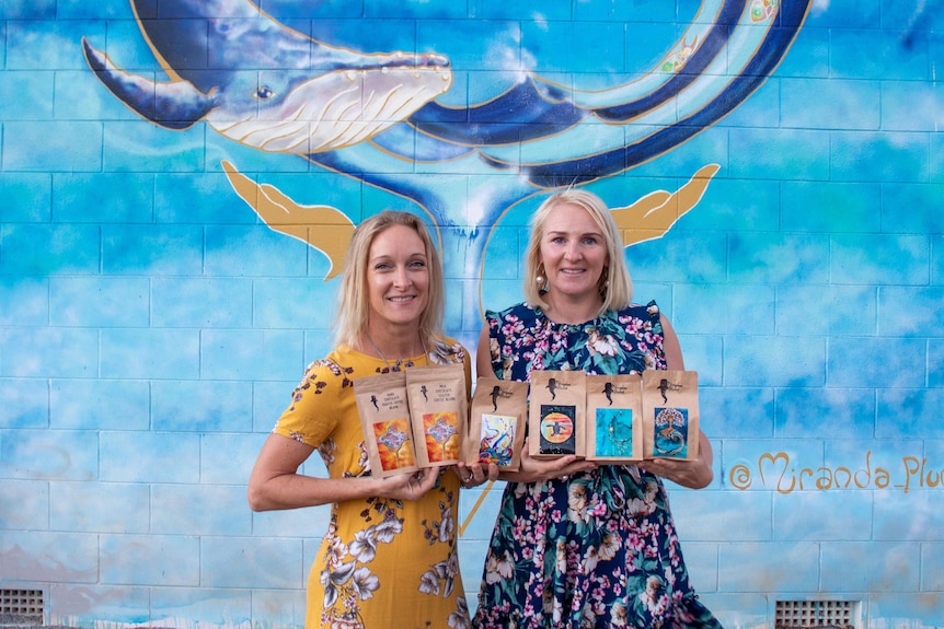 Two women wearing colourful dresses stand holding coffee bags in front of a mural