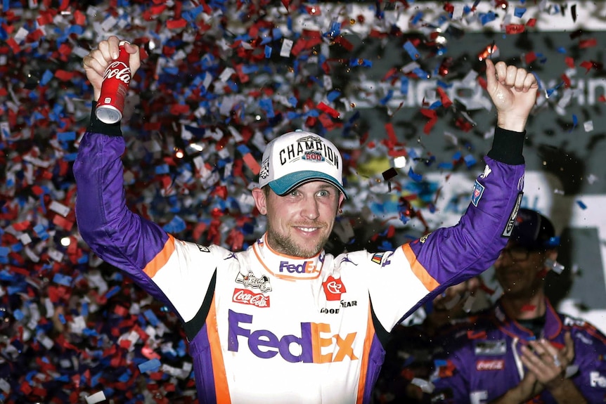 A NASCAR driver raises his arms in celebration, as he wears a cap saying "Daytona 500 champion".