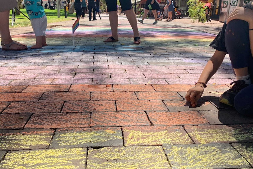 A brick mall colored using chalk in rainbow colors with people walking on the mall.