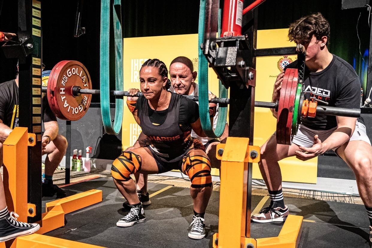 Lily squats with heavy bar - looks strained with weights. Men stand around her helping with weight. 