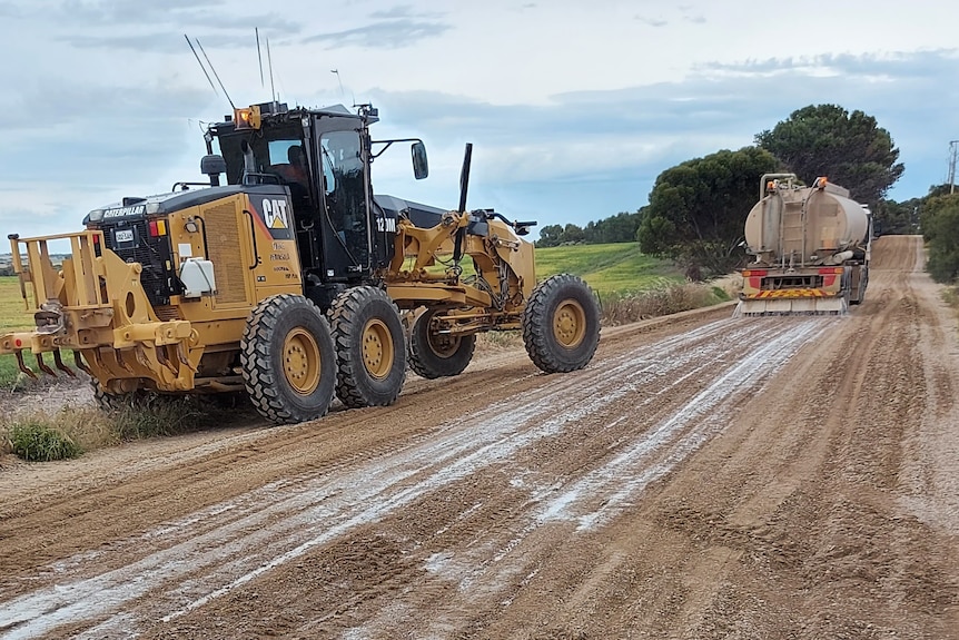 A yellow tractor drives behind a water truck applying a liquid coating onto a dirt road.