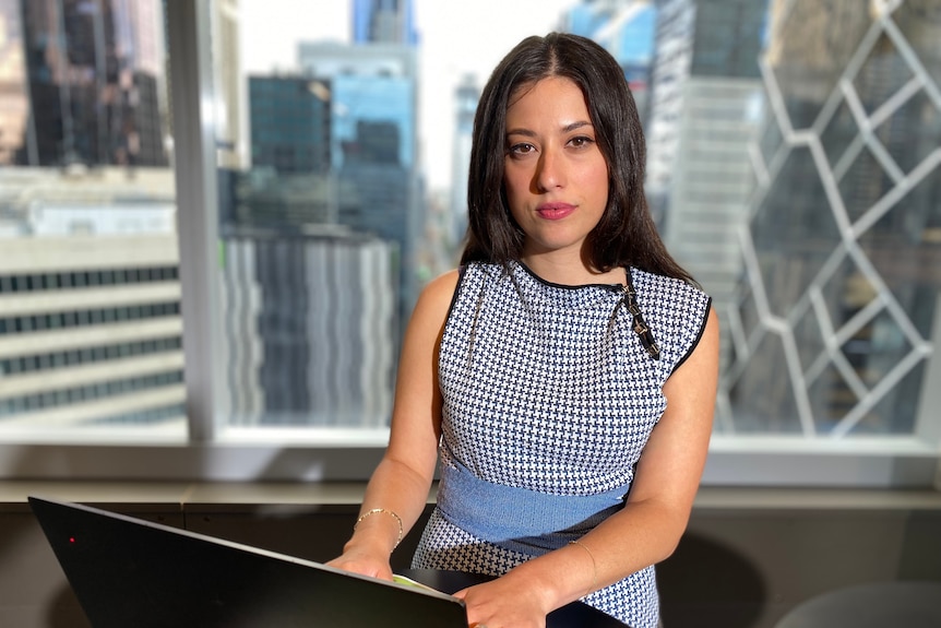 Zoe looks at the camera while typing on a laptop. The city skyline is visible behind her.