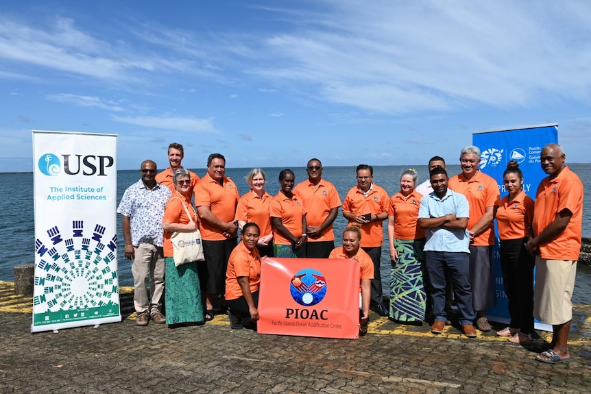 About a dozen Pacific Island scientists in organge shirts branded with PIOAC at the edge of Suva harbour
