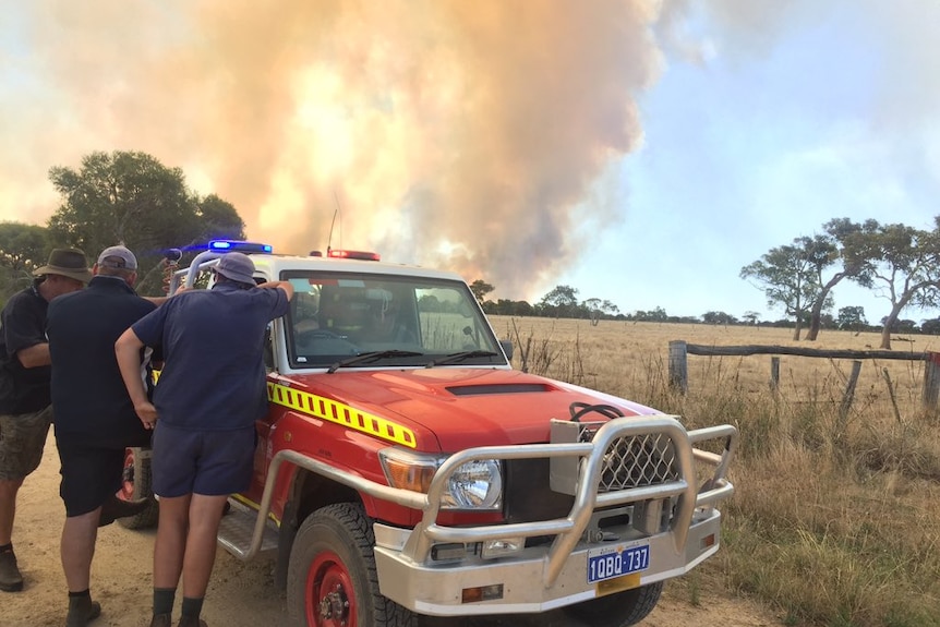 Three men stand next to a fire vehicle alongside a paddock as smoke rises in the distance.