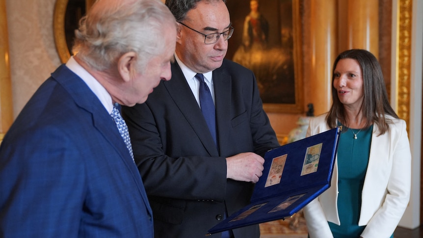 King Charles looks at a book with bank notes in it being held by a man with a woman standing in the background. 