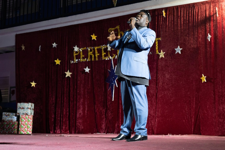 Mgr Lawrence Rolle sur scène vêtu d'un costume bleu foncé et tenant un microphone. 