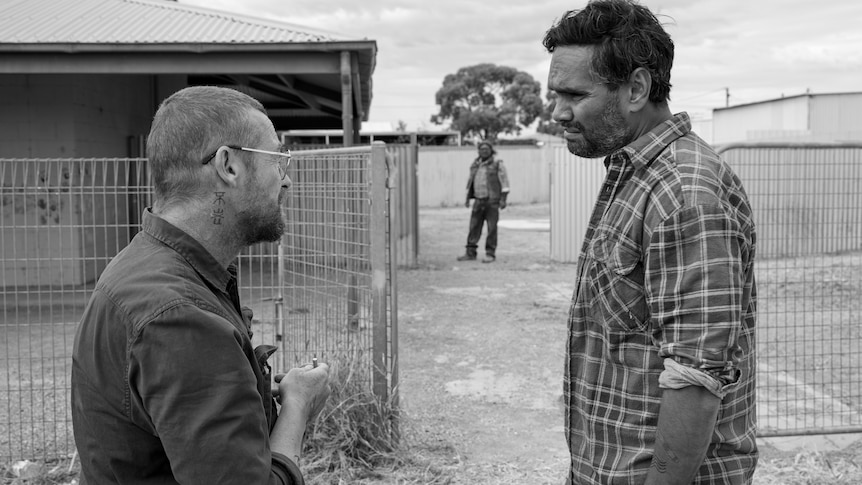 A black and white photo of two men, one Indigenous, standing opposite each other. A third man is seen in the background.