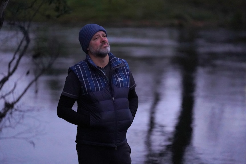 Man in beanie and puffer vest jacket stands looking solemn next to a lake 