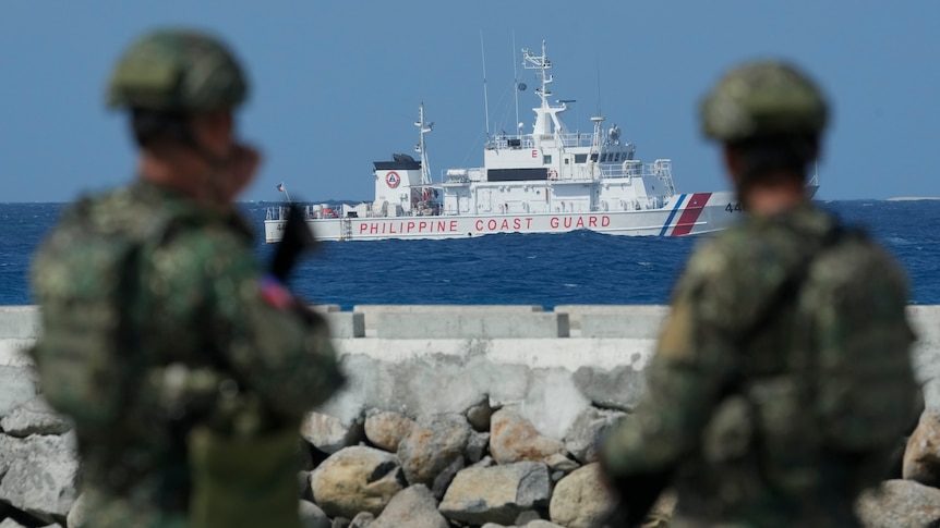 Two soldiers in camouflage look at a Philippine Coast Guard vessel sailing on open water 