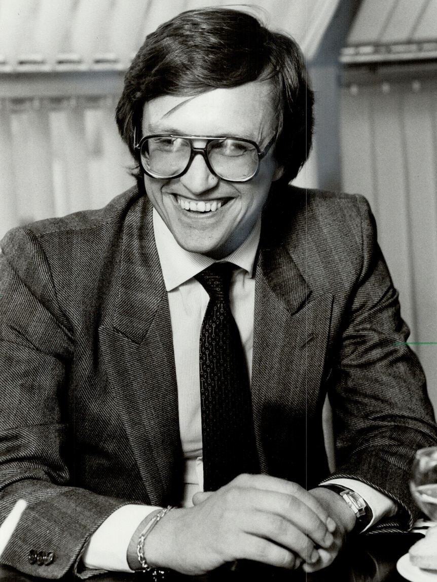 Black-and-white portrait shows Maurizio Gucci as a young man wearing aviator glasses and a suit, smiling widely, hands clasped