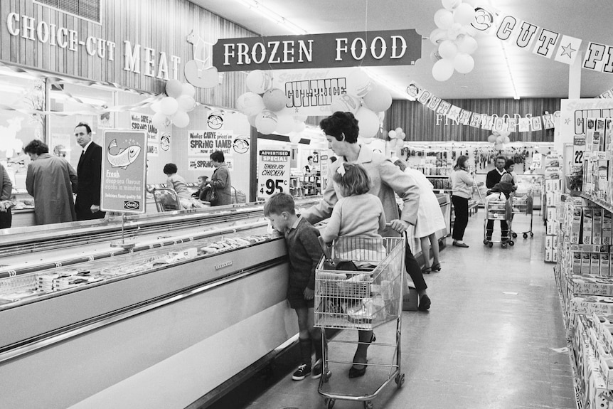 A 1960s black and white photo of a supermarket where a woman and two children look in a frozen food section