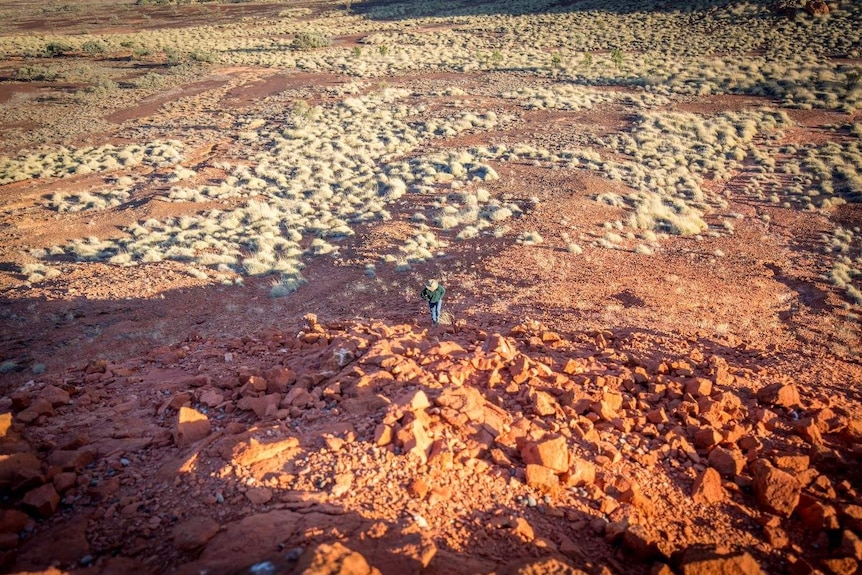 A man stands at the base of a rocky hill, with acres of arid grasslands spreading out behind him.